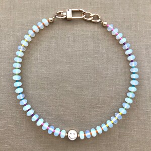 Pode incluir: Um colar de contas de opala branca com um fecho dourado e um pingente de carinha sorridente dourado. As contas s&atilde;o separadas por fio colorido.