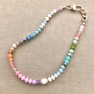 Pode incluir: Um colar de contas de arco-&iacute;ris com um fecho dourado e uma grande p&eacute;rola. O colar &eacute; feito com v&aacute;rias pedras semipreciosas e apresenta um degrad&eacute; de cores que vai do rosa ao azul.