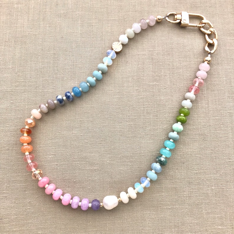 Pode incluir: Um colar de contas de arco-&iacute;ris com um fecho dourado e uma grande p&eacute;rola. O colar &eacute; feito com v&aacute;rias pedras semipreciosas e apresenta um degrad&eacute; de cores que vai do rosa ao azul.