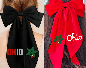 May include: Two large hair bows, one black and one red, each featuring long fabric tails. The black bow displays the text OHIO in red and white letters with a green buckeye leaf graphic. The red bow features the word Ohio in white script.