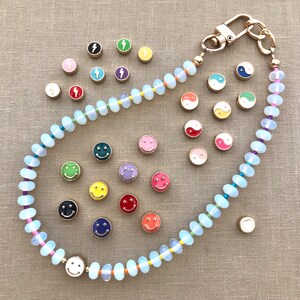 Pode incluir: Um colar de contas brancas com um fecho dourado e v&aacute;rios pingentes de esmalte coloridos, incluindo carinhas sorridentes, raios e s&iacute;mbolos yin-yang.
