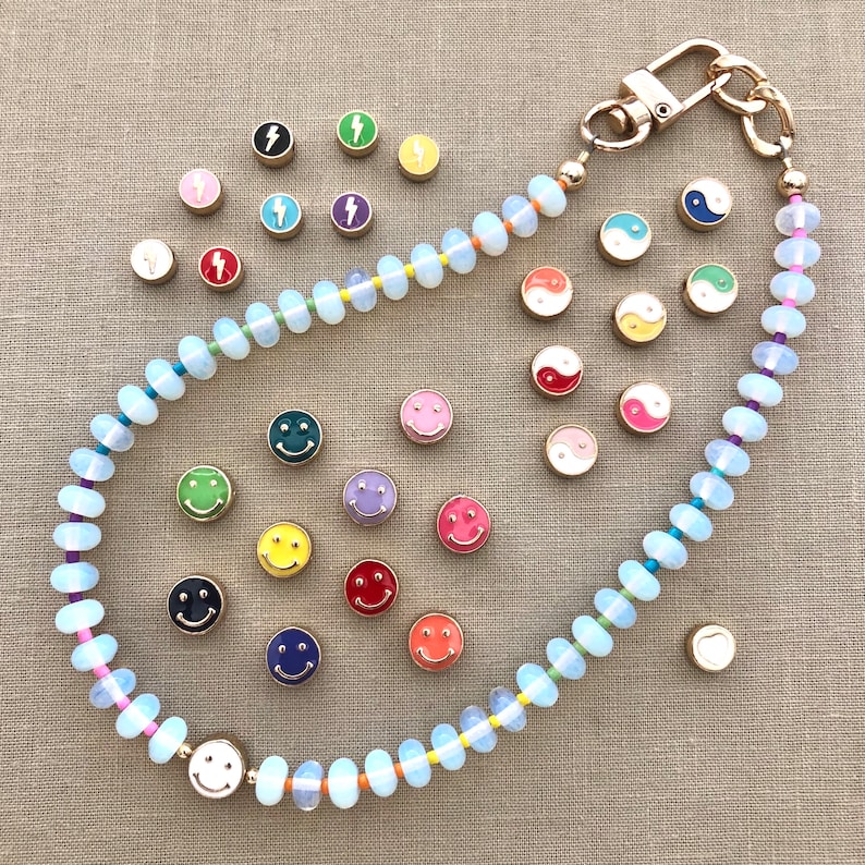 Pode incluir: Um colar de contas brancas com um fecho dourado e v&aacute;rios pingentes de esmalte coloridos, incluindo carinhas sorridentes, raios e s&iacute;mbolos yin-yang.