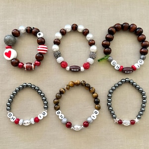 May include: Six different bracelets with various beads and charms. The bracelets feature red, white, brown, gray, black, and gold beads. Some bracelets have charms such as a football, a star, and a heart. The bracelets spell out "OHIO".