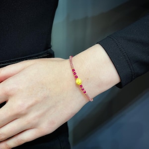 Peut inclure: Un bracelet délicat avec une perle jaune centrale, flanquée de petites perles rouges rondes. Le fil du bracelet est rose pâle, et le design est simple et élégant. Le bracelet est porté au poignet.