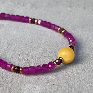 Puede incluir: Pulsera de cuentas con pequeñas cuentas cuadradas magenta, intercaladas con detalles dorados y cuentas rojas oscuras. Una gran cuenta redonda amarilla es el punto focal.