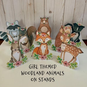 May include: A set of eight woodland animal figurines on stands. The animals are painted in watercolor style and include a fox, deer, bear, raccoon, squirrel, bunny, and two skunks. The stands are decorated with flowers and greenery. The text "GIRL THEMED WOODLAND ANIMALS ON STANDS" is printed below the figurines.