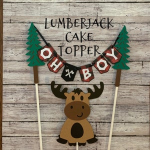 May include: Lumberjack cake topper with a red and black plaid banner that says "Oh Boy", a moose, and two green pine trees.