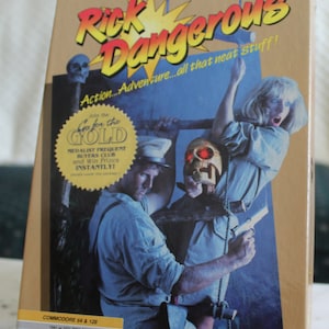 Puede incluir: Una caja para el juego de computadora Rick Dangerous para Commodore 64 y 128. La caja presenta un gráfico de explosión amarillo y rojo con el título "Rick Dangerous" en letras rojas y negritas. La caja también presenta una foto de un hombre y una mujer en un entorno de jungla. El hombre sostiene un arma y la mujer está colgada de una cuerda. El texto "Action...Adventure...all that neat stuff!" está impreso en la caja.