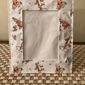 May include: A white fabric picture frame featuring a repeating pattern of Winnie the Pooh characters, including Pooh Bear, Tigger, and Piglet. The frame has a rectangular opening for a photo and is bordered with a stitched edge.