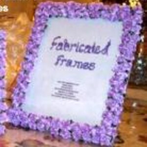 Op de afbeelding: Een rechthoekige fotolijst versierd met talloze kleine, lichtpaarse stoffen bloemen. Het midden van de lijst bevat een witte inzet met de tekst "Fabricated Frames" in een donkerpaars cursief lettertype, gevolgd door kleinere gedrukte tekst.