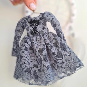 May include: A miniature grey lace dress with long sleeves and a flared skirt. The dress has a black floral embellishment at the bodice. The delicate lace fabric is visible throughout the dress, creating a textured appearance. The dress is held up by a small hanger.