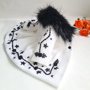 May include: A white beanie hat with black floral embroidery and a black faux fur pom-pom. The hat features a detailed floral design around the brim and on the side. A small cluster of orange roses is in the background.