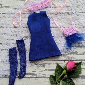 Pode incluir: Um vestido azul sem mangas, um colar de renda rosa, meias de renda azul e uma bolsa de crochê rosa com um detalhe de pena azul. Uma única rosa rosa com folhas verdes está sobre uma superfície de madeira branca.