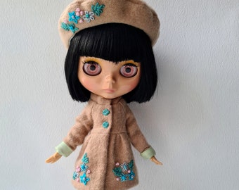 Puede incluir: Una muñeca con un abrigo y boina color beige, ambos adornados con bordados florales azules y blancos. La muñeca tiene cabello negro, zapatos rosas y una pequeña silla blanca. El abrigo tiene puños verdes y tres botones.