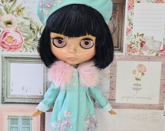 May include: A doll with black hair and large eyes wearing a mint green coat with floral embroidery, a matching beret, and a pink fur collar. The doll is also wearing white lace stockings and is posed in front of a decorative background.