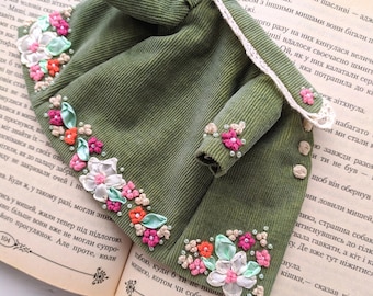 May include: An olive green miniature jacket with floral embroidery in pink, white, and orange. The jacket features a corduroy texture and is embellished with small beads. It is displayed open on the pages of an old book.