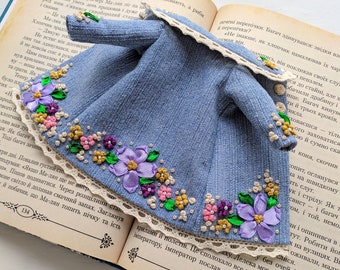 Könnte beinhalten: Miniatur-Jeansjacke mit floralen Stickereien in Lila, Rosa und Gelb. Die Jacke hat eine Spitzenborte und liegt auf einem offenen Buch.