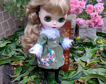 Puede incluir: Una muñeca con cabello rubio que lleva un abrigo de pana verde con ribete de piel blanca y bordado floral rosa. La muñeca está de pie sobre una cama de hojas verdes.