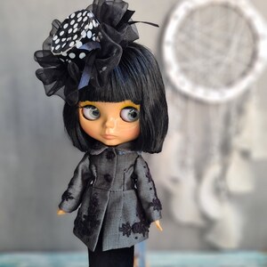 May include: A black and white polka dot hat with a black bow and veil on a doll with black hair, wearing a gray and black jacket and a black skirt. The doll is standing on a blue stand.