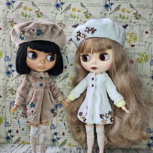 Può includere: Due bambole con i capelli lunghi che indossano abiti bianchi e marroni con ricami floreali. Indossano berretti e calze di pizzo bianco. Le bambole sono in piedi su una superficie di legno bianco.