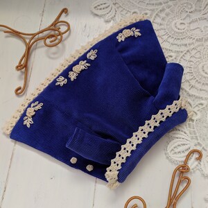 May include: A blue corduroy jacket with white lace trim and embroidered flowers. The jacket has a button closure and a long sleeve.