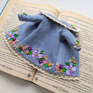 以下が含まれることがあります： 紫、ピンク、黄色の花の刺繍が施されたミニチュアデニムジャケット。 ジャケットはレースのトリム付きで、開いた本の上に置かれています。