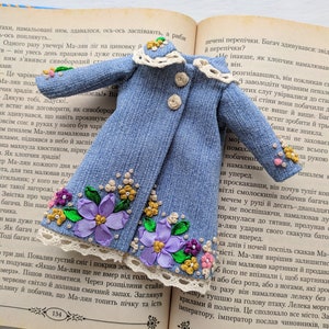 以下が含まれることがあります： 刺繍された紫の花、黄色のアクセント、白いレースのトリムで飾られた、ミニチュアのデニムコート。小さなコートには、ベージュのボタンが3つと白いレースの襟が付いています。開いた本の上に置かれています。