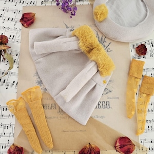 May include: A light gray felted hat with a mustard yellow faux fur trim, a matching light gray beret with a mustard yellow pom-pom, and two pairs of mustard yellow textured stockings, all arranged on a sheet of vintage musical notation.