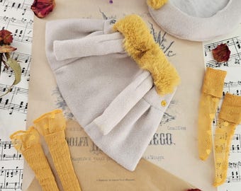Op de afbeelding: Een lichtgrijze vilten hoed met een mosterdgele imitatiebontrand, een bijpassende lichtgrijze baret met een mosterdgele pompon en twee paar mosterdgele gestructureerde kousen, alles gerangschikt op een blad met oude muzieknoten.