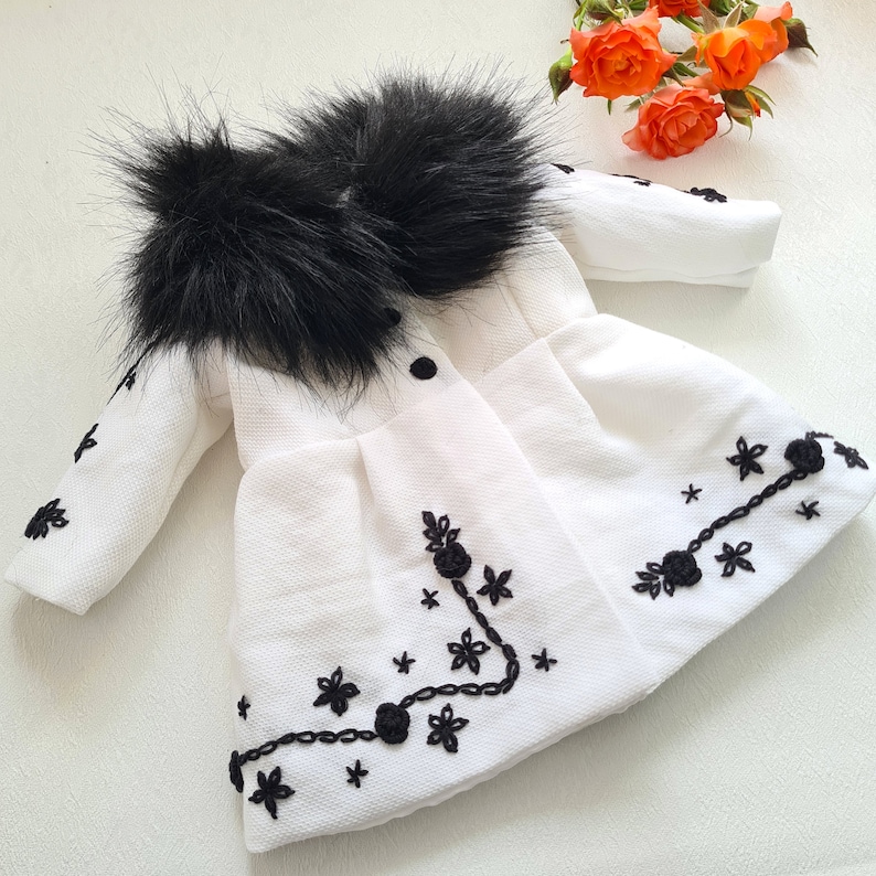 May include: A white faux fur coat with a black fur collar and black embroidered floral details.