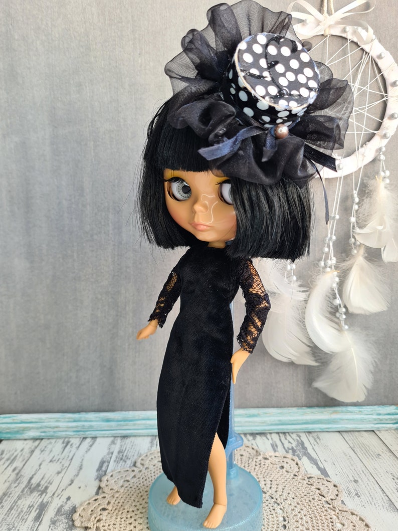 May include: A black and white polka dot top hat with a black tulle bow and a black velvet dress with lace sleeves. The doll has black hair and blue eyes.