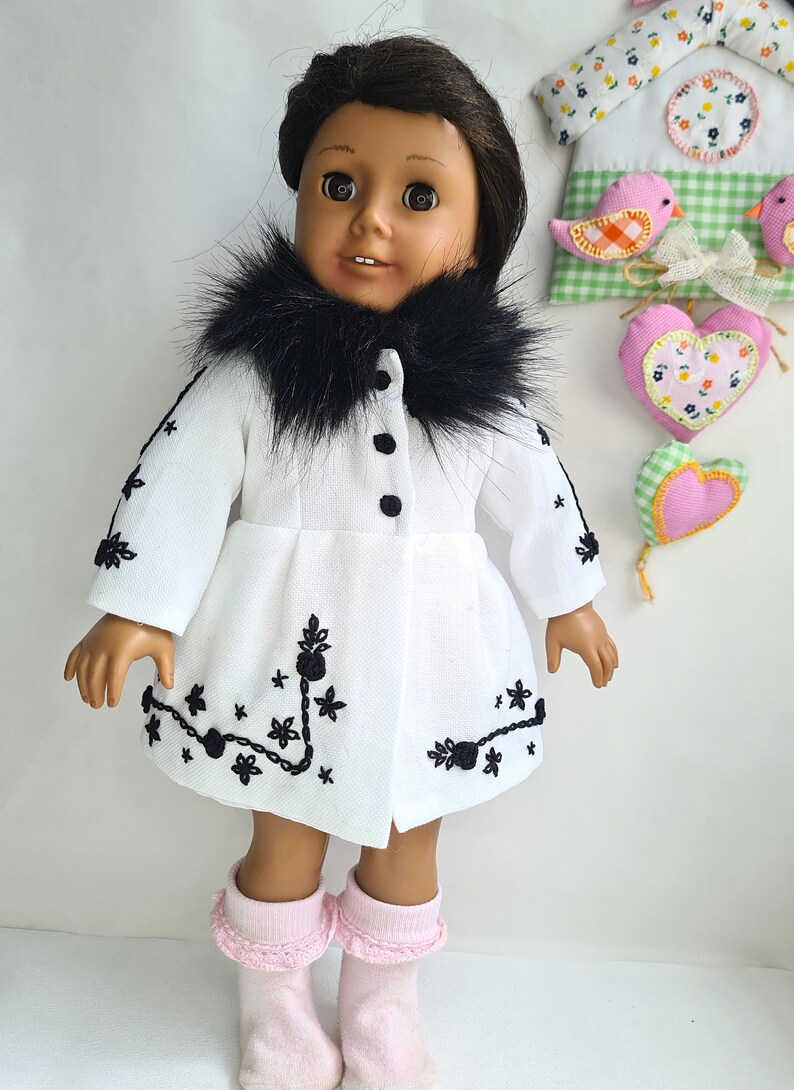 May include: A white doll wearing a white coat with a black faux fur collar and black embroidery. The doll has brown skin and black hair. The doll is wearing pink socks.