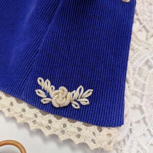 May include: A close-up of a blue corduroy fabric with a white embroidered flower design. The fabric has a white lace trim.