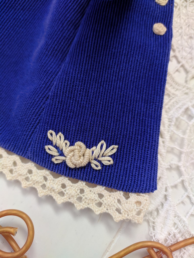 May include: A close-up of a blue corduroy fabric with a white embroidered flower design. The fabric has a white lace trim.