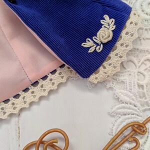 May include: A blue corduroy jacket with a white embroidered floral design on the sleeve. The jacket has a pink lining and a white lace trim.