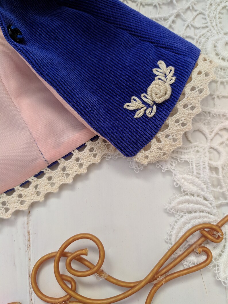May include: A blue corduroy jacket with a white embroidered floral design on the sleeve. The jacket has a pink lining and a white lace trim.
