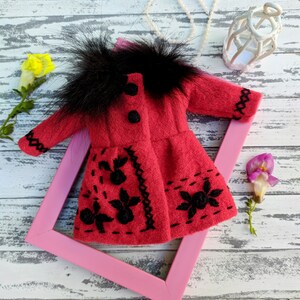 May include: A red felt doll coat with black embroidery and a black faux fur collar. The coat is displayed on a pink frame against a white wooden background.