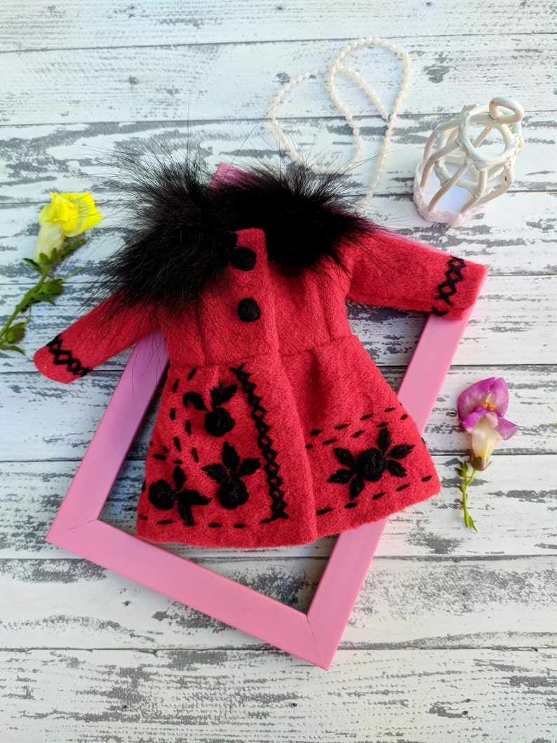 May include: A red felt doll coat with black embroidery and a black faux fur collar. The coat is displayed on a pink frame against a white wooden background.