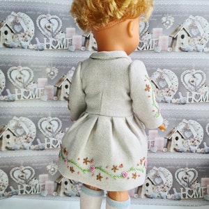 K&ouml;nnte beinhalten: Eine Puppe tr&auml;gt ein hellgraues Kleid mit gestickten Blumen. Das Kleid hat einen Faltenrock und einen Kragen. Die Puppe tr&auml;gt wei&szlig;e Socken mit blauen R&auml;ndern.