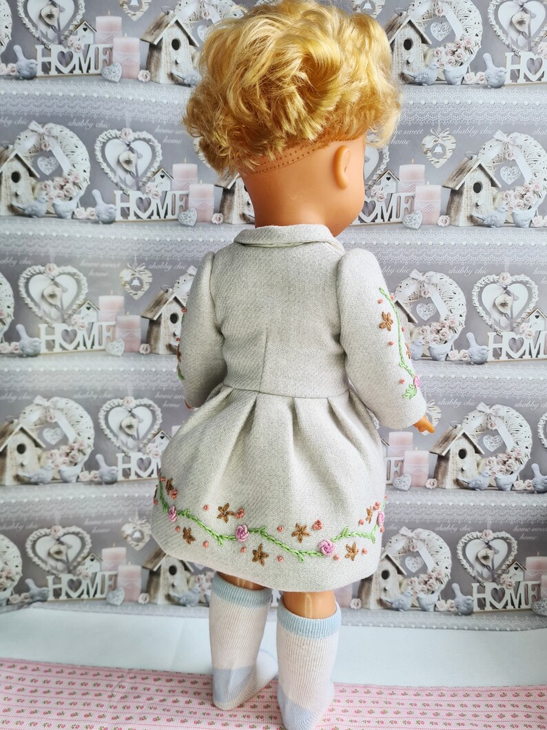 K&ouml;nnte beinhalten: Eine Puppe tr&auml;gt ein hellgraues Kleid mit gestickten Blumen. Das Kleid hat einen Faltenrock und einen Kragen. Die Puppe tr&auml;gt wei&szlig;e Socken mit blauen R&auml;ndern.