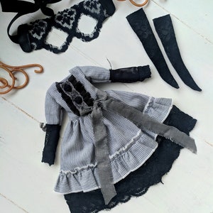 May include: A doll dress with a black and white striped pattern, black lace trim, and a grey ribbon. The image also includes a black lace eye mask and two black lace arm cuffs. The items are arranged on a white wooden surface.