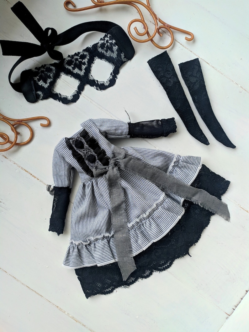 May include: A doll dress with a black and white striped pattern, black lace trim, and a grey ribbon. The image also includes a black lace eye mask and two black lace arm cuffs. The items are arranged on a white wooden surface.