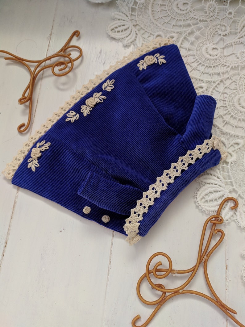 May include: A blue corduroy jacket with white lace trim and embroidered flowers. The jacket has a button closure and a long sleeve.