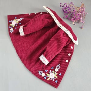May include: A burgundy corduroy dress with long sleeves and a white lace collar. The dress features floral embroidery in pink, blue, and white, and white buttons. The cuffs are trimmed with a floral patterned fabric.