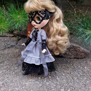 May include: A doll with blonde hair and green eyes wearing a black lace mask and a grey and white striped dress with black lace trim. The doll is posed outdoors on a textured surface.