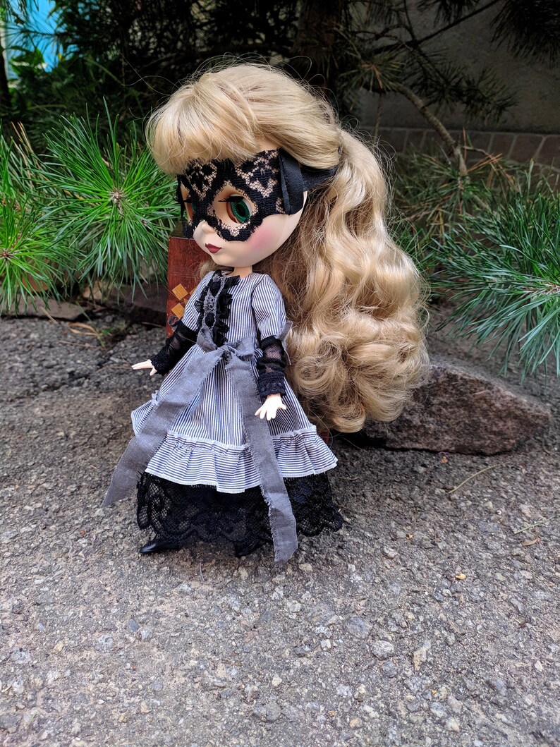 May include: A doll with blonde hair and green eyes wearing a black lace mask and a grey and white striped dress with black lace trim. The doll is posed outdoors on a textured surface.