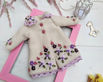 May include: A cream-colored doll coat with floral embroidery in purple, pink, and green. The coat features a scalloped collar and hem, with matching buttons. It is displayed on a pink picture frame, with floral and decorative elements in the background.