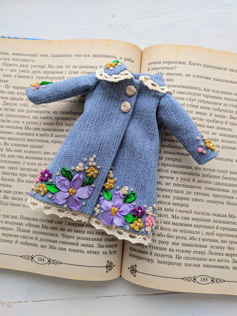 以下が含まれることがあります： 刺繍された紫の花、黄色のアクセント、白いレースのトリムで飾られた、ミニチュアのデニムコート。小さなコートには、ベージュのボタンが3つと白いレースの襟が付いています。開いた本の上に置かれています。