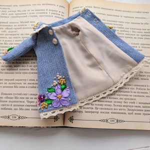 以下が含まれることがあります： 開いた本の上に置かれた、花の刺繍とレースのトリムが付いたミニチュアの青いデニムジャケット。 ジャケットは、明るいベージュの裏地と3つの小さなボタンが特徴です。