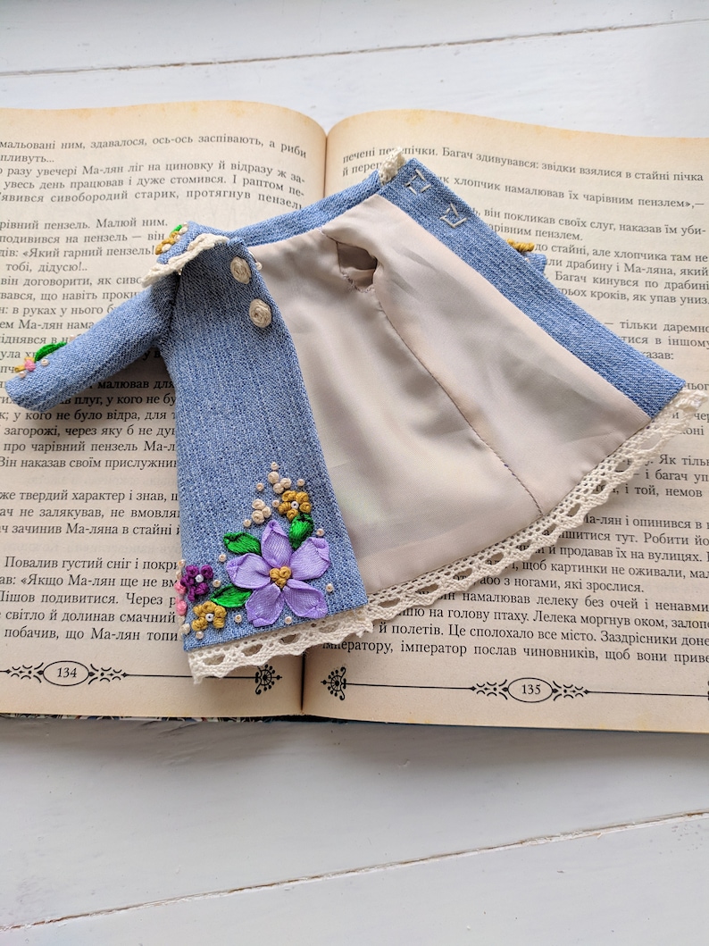 以下が含まれることがあります： 開いた本の上に置かれた、花の刺繍とレースのトリムが付いたミニチュアの青いデニムジャケット。 ジャケットは、明るいベージュの裏地と3つの小さなボタンが特徴です。